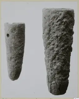 Two tapering bronze tools side by side, the smaller one with a small hole near its top, the larger one with a pitted, knobbly surface.