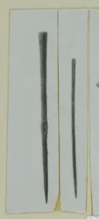 Two long, thin bronze tools with pointed ends, shown upright side by side on a pale card.