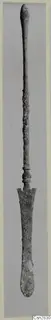 Long bronze implement with a slender shaft and a flat spatula-like blade at the lower end, photographed upright on a grey backdrop.