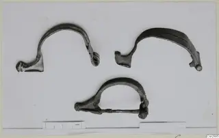 Three bronze bow-shaped personal adornments on a pale background, with a scale bar along the bottom edge.