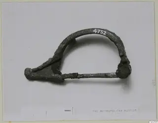 Bronze bow-shaped brooch with a long pin, photographed flat beside a scale bar