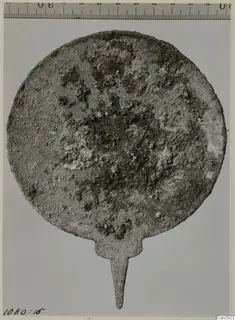 Corroded round metal disc with a pointed tang, photographed flat with a ruler along the top edge