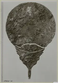 Corroded metal disc with a handle ending in a human face, photographed beside a scale bar