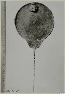 Corroded round metal disc with a long thin stem, shown beside a measuring rule.