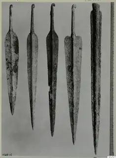 Five corroded metal blades with pointed tips and narrow tangs, photographed side by side against a grey backdrop.