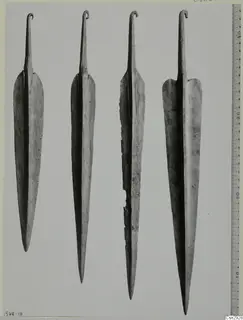 Four long metal blades with hooked tangs at the top, photographed side by side beside a measuring ruler