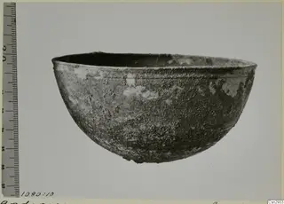 Corroded bowl with a rounded body and a thick rim, photographed beside a scale ruler
