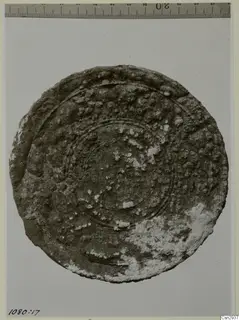 Round, heavily corroded object with concentric ridges and a pale crust, shown against a grey background with a scale above.