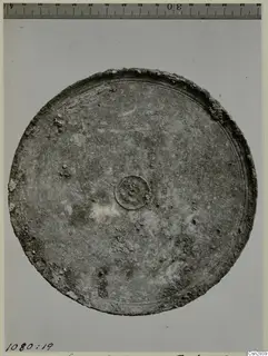 Round metal object with a raised rim and small central boss, photographed beside a scale ruler