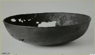 Dark grey bowl with a rounded body and rim, with holes broken through its wall, photographed on a plain background.