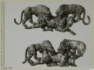 Two corroded openwork metal figures, each showing two maned cat-like animals with curled tails, shown one above the other