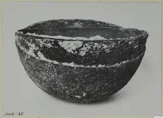 Deep, rounded bowl with a rough, encrusted dark surface and a wide rim, photographed on a pale background.