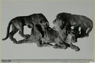 Dark, corroded metal figure group of animals, with a lion on the left, photographed beside a millimetre scale ruler