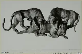 Dark metal figure of two maned animals attacking a third animal below, shown with a centimetre scale beneath
