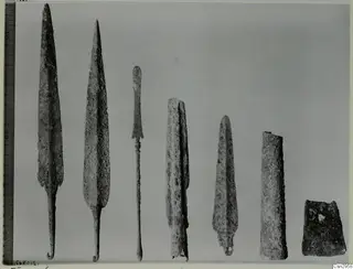 Row of seven corroded metal objects, including two long pointed blades, laid out on a grey background