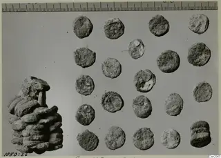 Corroded round metal discs, probably coins, laid out in rows beside a stack of similar discs, with a ruler along the top.