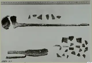Corroded iron objects and small dark fragments laid out on a pale ground, with a centimetre ruler along the top edge