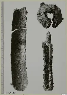 Three corroded metal objects on a pale background: a long flat strip broken in two, a ring with a central hole, and a pointed rod.