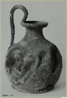 Pottery vessel with a single loop handle rising from its shoulder to the rim, its surface crusted with soil