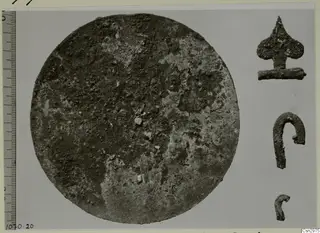 Corroded circular metal disc, with three small corroded metal objects to its right and a measuring scale along the left edge