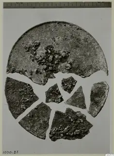Broken fragments of a corroded circular disc on a pale background, with a measuring ruler along the top edge