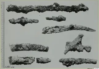 Nine corroded metal fragments on a pale background, with a measuring scale along the left edge.