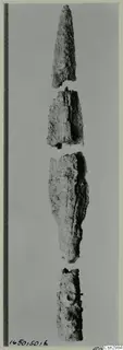 Four corroded metal fragments stacked vertically, the top one tapering to a point, with a measuring scale along the left edge.