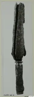 Long corroded metal object with a split, blade-like upper end above a ring collar, shown beside a measuring scale.