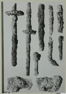 Corroded metal objects before conservation, including two sword-like pieces with cross-guards, laid out beside a scale bar