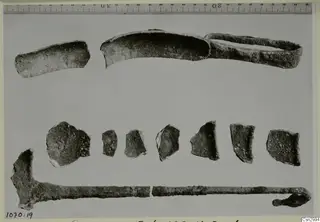 Corroded metal fragments laid out in rows beside a ruler, including a curved strip and a long rod with a hooked end.