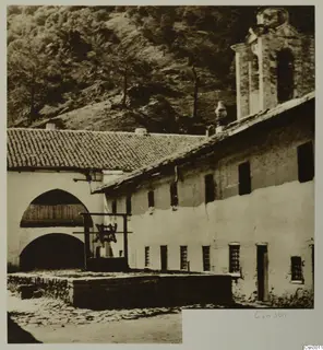 Whitewashed building with a bell hung in a wooden frame at Machairas, Troodos, with a wooded hillside behind.
