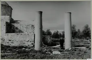 Man in a dark suit standing between two stone columns beside a ruined building, Paphos
