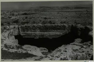 Dark rock-cut opening under an overhanging rock ledge in open scrubby ground, with low hills behind