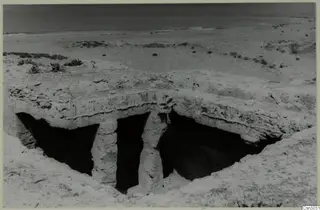 Flat-roofed rock structure resting on two pillars, with dark openings beneath it, on sandy ground above a calm sea.