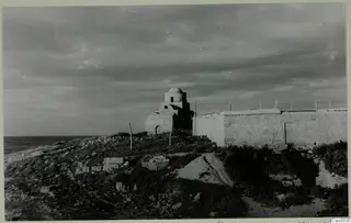 Small domed building beside a long whitewashed wall and wire fence on a rocky coastal rise