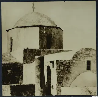 Whitewashed domed church with a cross on top and a small window, with a stone wall and arched doorway in front.