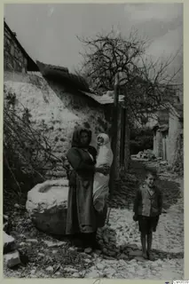 Woman holding a baby and a boy standing on a cobbled lane beside a large stone basin and a stone wall, Paphos
