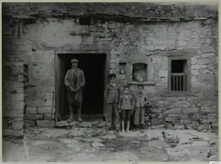 Man and three boys standing at the doorway of a stone building, with a bird on the paved ground in front