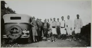Group of people standing beside an old car on a dirt road, with a large pot and bottles near their feet