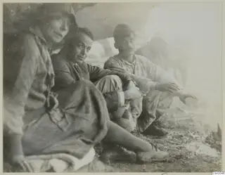 Four people sitting and resting on dry ground at a site, one in a hat and one in a long skirt, in a bright hazy setting.