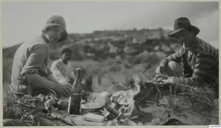 Picnic on a grassy hillside: a woman and a man in hats with a glass bottle, food and paper wrappings in the foreground