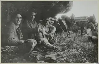 People and a loaded donkey resting under a large rock overhang, with grass and low plants in the foreground.