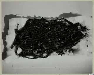 Dark woven basket of twigs or rods lying on a pale surface, photographed from above.