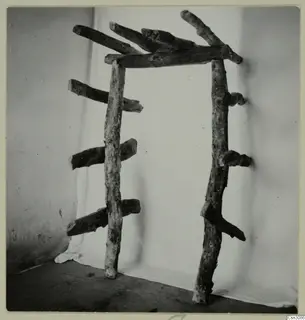 Wooden frame of rough, bark-covered branches standing on a floor against a white cloth, from the Cypriote Mining group.