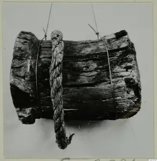 Weathered wooden cylinder with a braided rope hanging across it, tied with thin cords against a plain grey background