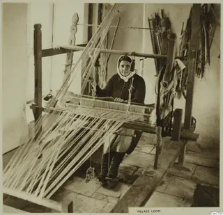 Woman in a headscarf seated at a wooden village loom, with long warp threads stretched across the frame