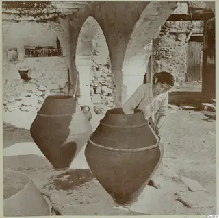Woman leaning over a large earthenware wine jar in a stone-walled courtyard, with a second jar beside her