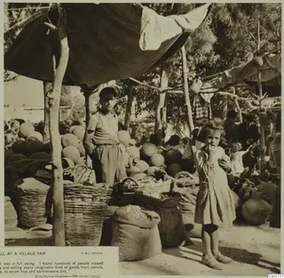 Potter's stall at a village fair: round pots, baskets and sacks under a canopy, with a boy and a small girl in front.