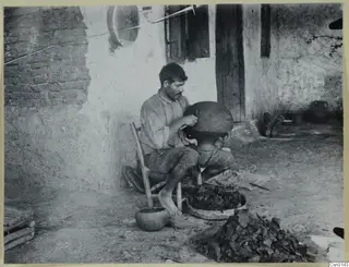 Man seated in a farmyard shaping a large round vessel with a wooden knife, Klirou