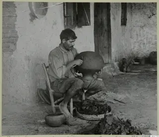 Man shaping a large clay vessel with a wooden knife, seated outside a whitewashed wall with a wooden door